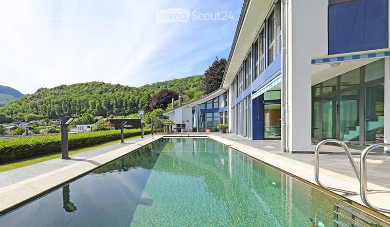 Modern outdoor area with a swimming pool, surrounded by a well-maintained lawn and a modern building with large windows and a blue accent wall. The background features a scenic view of green hills and trees.