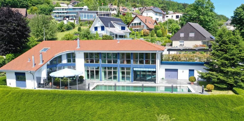 A modern two-story house with a red-tiled roof, large windows, and a swimming pool in the front yard. The house features a garage and is surrounded by lush greenery and neighboring houses.