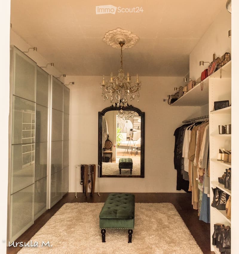 A well-lit, stylish room with a green tufted stool, a large ornate mirror, a wardrobe with hanging clothes, shelves with various items, and a chandelier hanging from the ceiling.