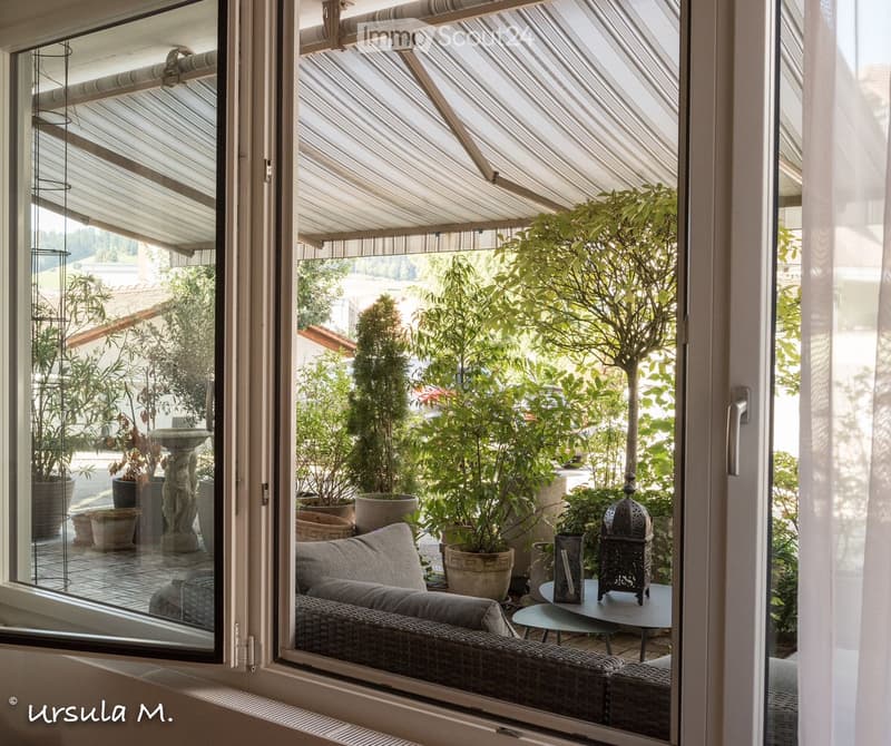 A spacious balcony with a wicker sofa, small round table, decorative table lamp, and several potted plants, enclosed by a striped awning.