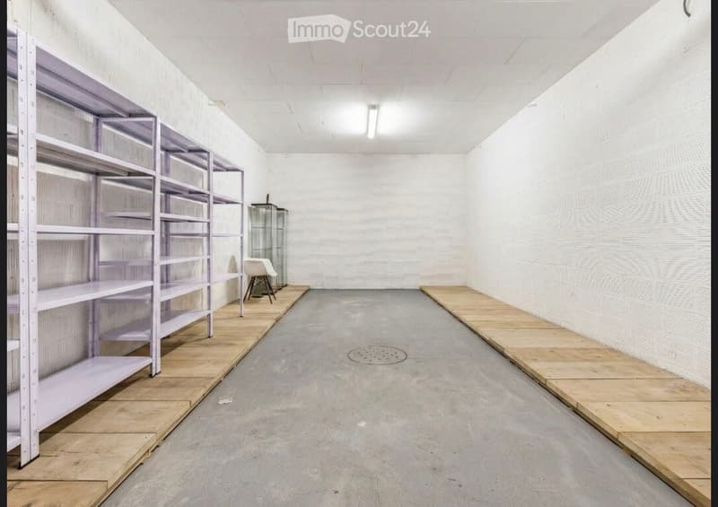 Empty room with white brick walls, concrete floor, metal shelving unit, and a small glass display case with a chair nearby.