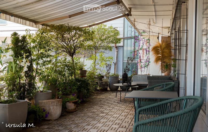 A covered outdoor terrace with a tiled floor, surrounded by potted plants and greenery, featuring outdoor furniture including tables and chairs.