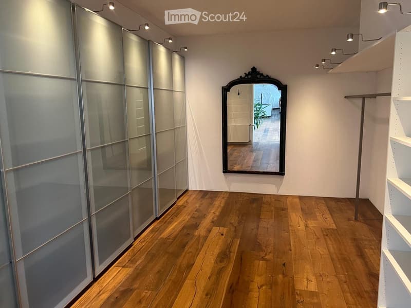 Empty hallway with wooden flooring, white walls, built-in closet with mirrored doors, and a decorative mirror on the wall.