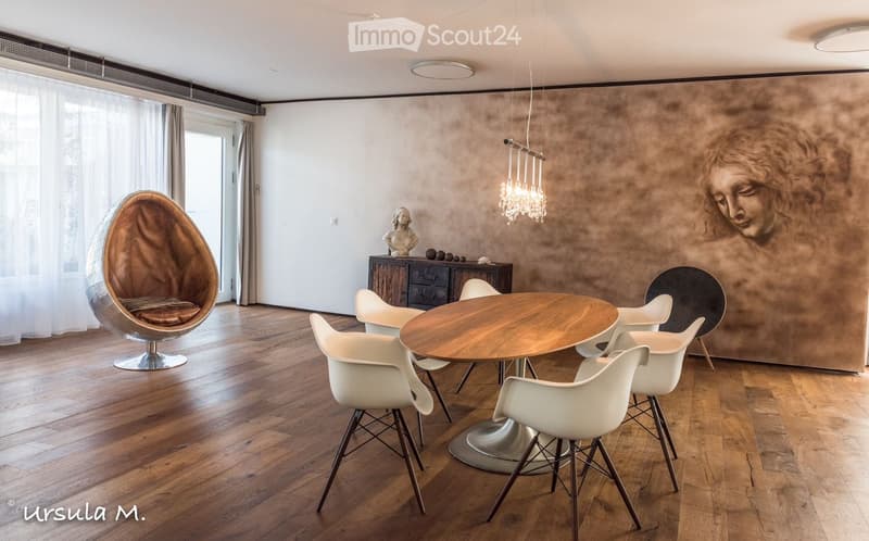 Modern dining area with a round wooden table surrounded by white chairs, a unique wicker egg chair, a wooden cabinet with a statue, and a chandelier hanging from the ceiling.
