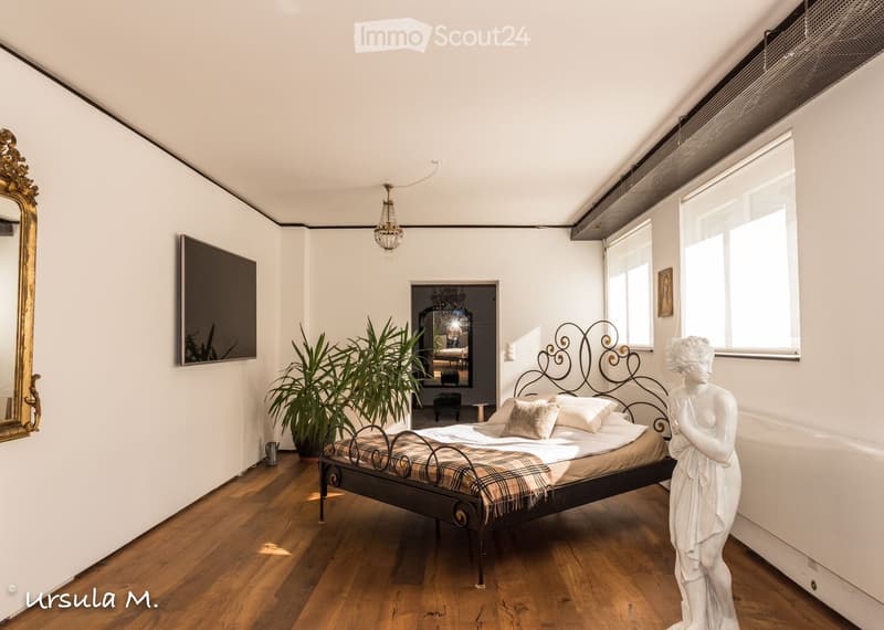 Elegant bedroom with wooden flooring, a large bed with a decorative metal headboard, a chandelier, a large mirror, a potted plant, and a white statue.
