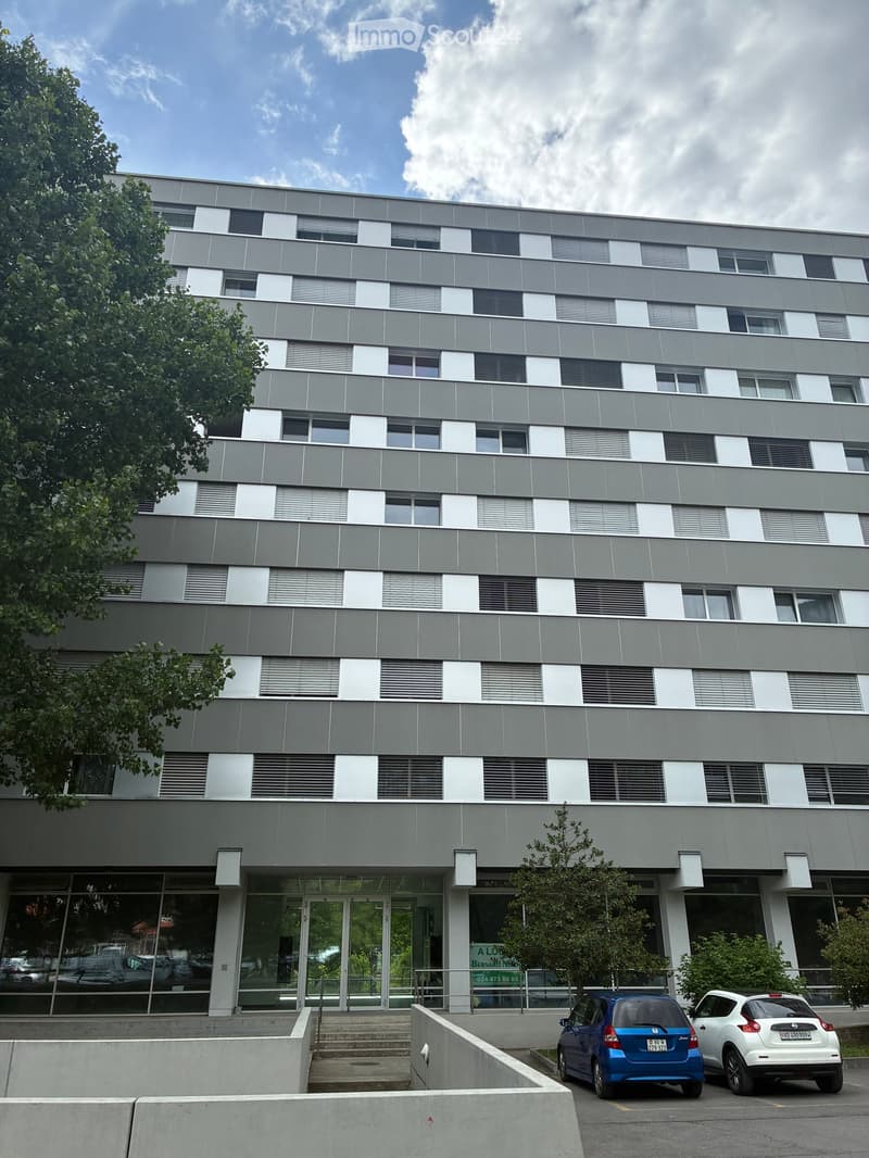 Modern multi-story building with grey and white facade, large windows, and a glass entrance. Two cars are parked in front.