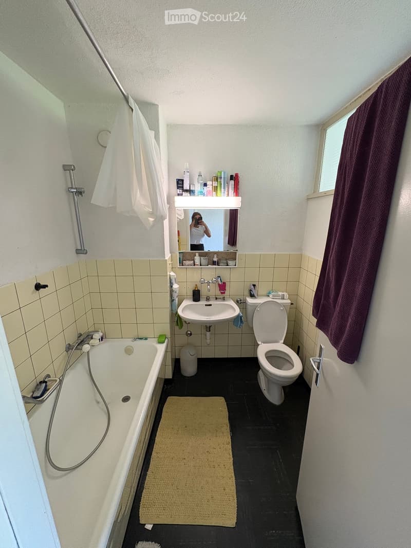 Small bathroom with a bathtub, toilet, sink, mirror, and a towel hanging on the wall. The floor is black, and there is a small beige bathmat in front of the sink.