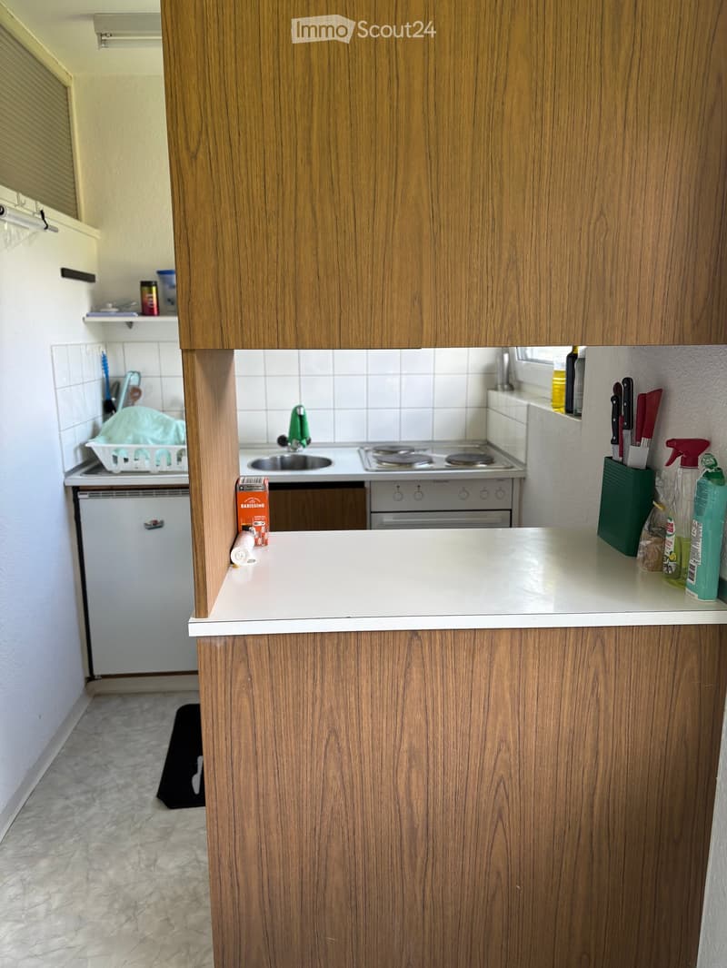 Modern kitchen with wooden cabinets, white countertop, sink, stove, oven, and dishwasher. Cleaning supplies and a knife holder are visible on the counter.