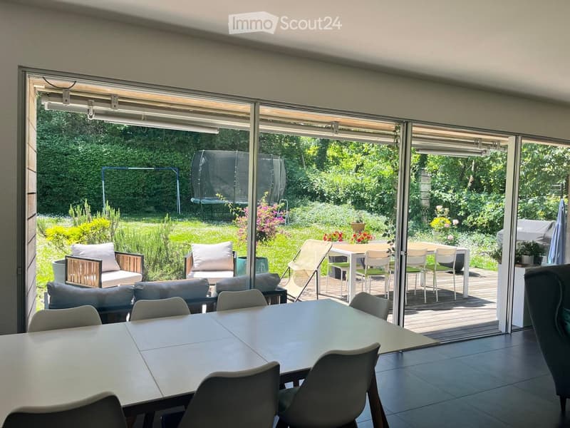 Bright room with large sliding glass doors opening to a garden, featuring a dining table with chairs, a sofa with pillows, and an ottoman.