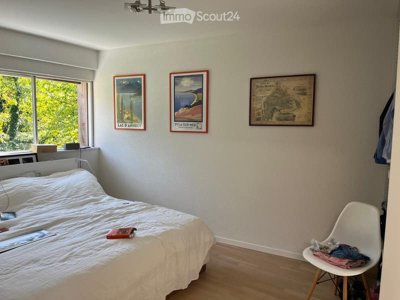 A well-lit bedroom with a white bed, a modern white chair, and three framed pictures on the wall. The room has wooden flooring and large windows with a view of greenery.