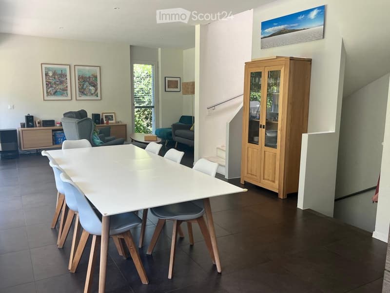 Modern dining area with a white dining table and chairs, a wooden cabinet, a sofa, an armchair, and a lamp. The room features large windows and artwork on the walls.