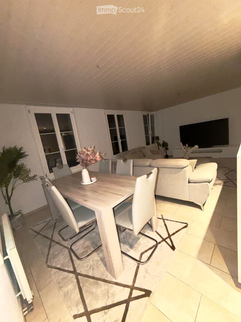Bright, modern dining area with a wooden dining table, white chairs, a white sofa, and a flat-screen TV in the background. The space features large windows and a tiled floor.