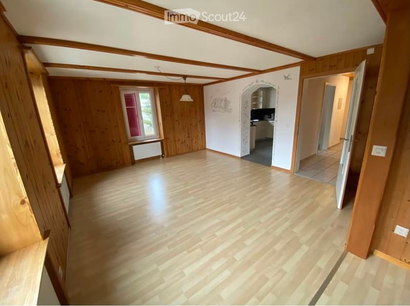 Empty room with wooden walls and flooring, large window with red shutters, visible kitchen area in the background.