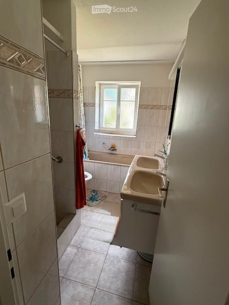 Bathroom with a double sink, bathtub, toilet, and a window above the bathtub. The walls are tiled, and there is a shower curtain visible.
