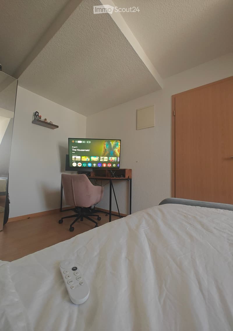 A modern bedroom with a neatly made bed, a pink upholstered chair, a desk with a television, and a remote control on the bed.
