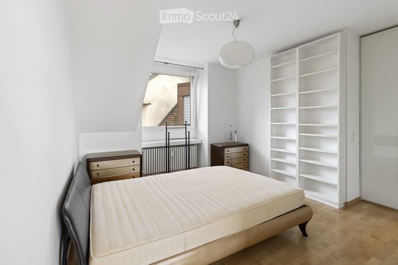 Furnished Room. A bedroom with a bed, nightstand, dresser, and shelves. The room has a white ceiling, walls, and wooden floor. The bed has a mattress and a headboard. The nightstand has a lamp and a bottle. The dresser has drawers. The shelf has a vase an