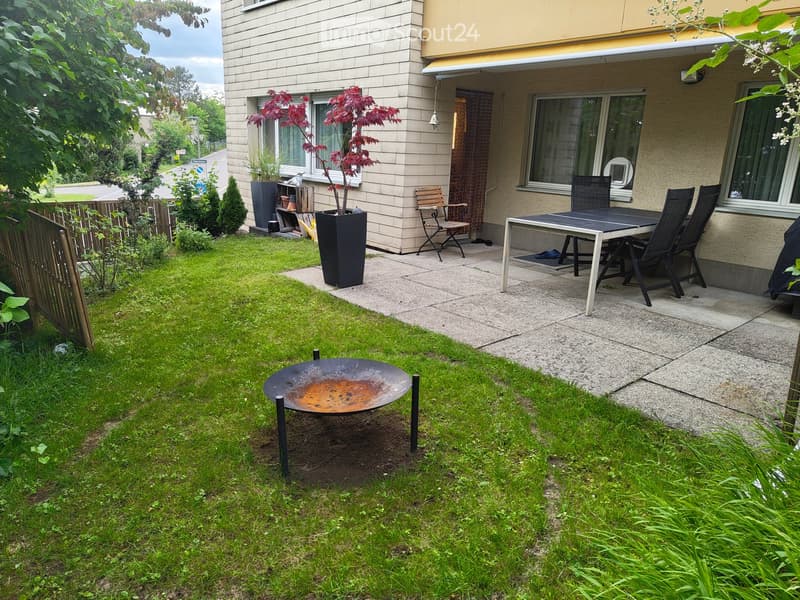 Garden with a well-maintained lawn, a round metal fire bowl, a patio area with a dining table and chairs, and potted plants near the house.