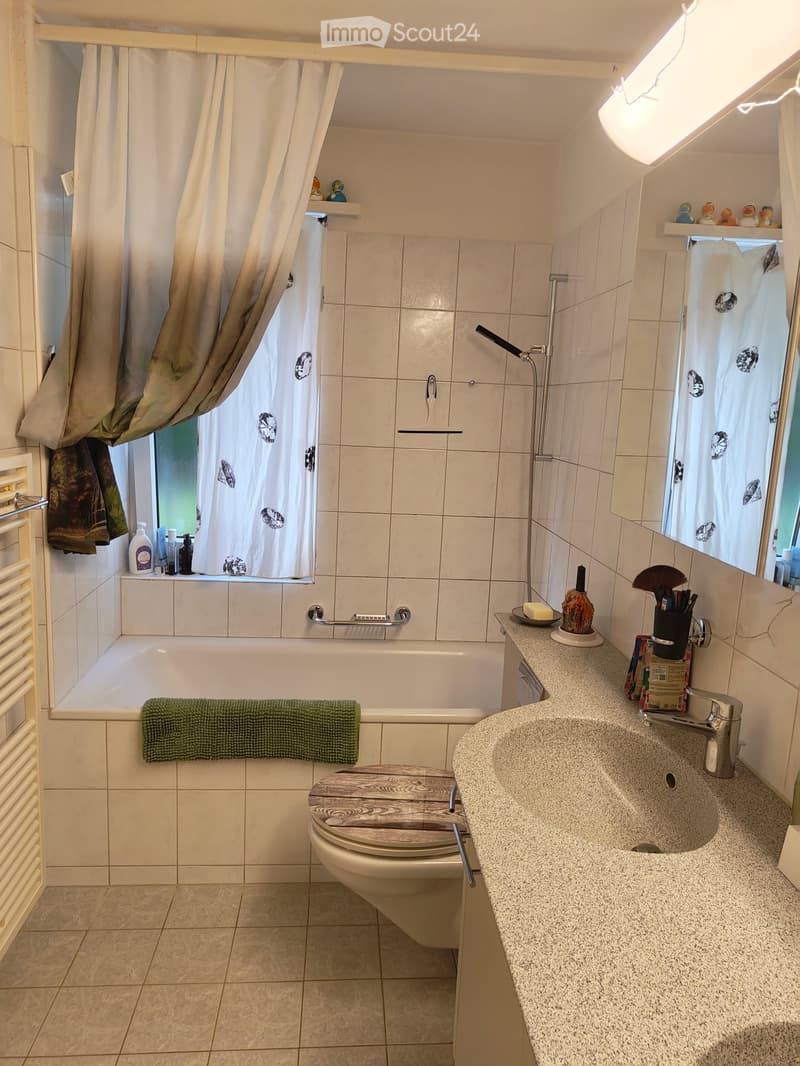 Bathroom with a bathtub, toilet, sink, and shower area. The sink has a modern faucet and a countertop with various toiletries. A towel hangs on the side of the bathtub, and a shower curtain is visible in the background.