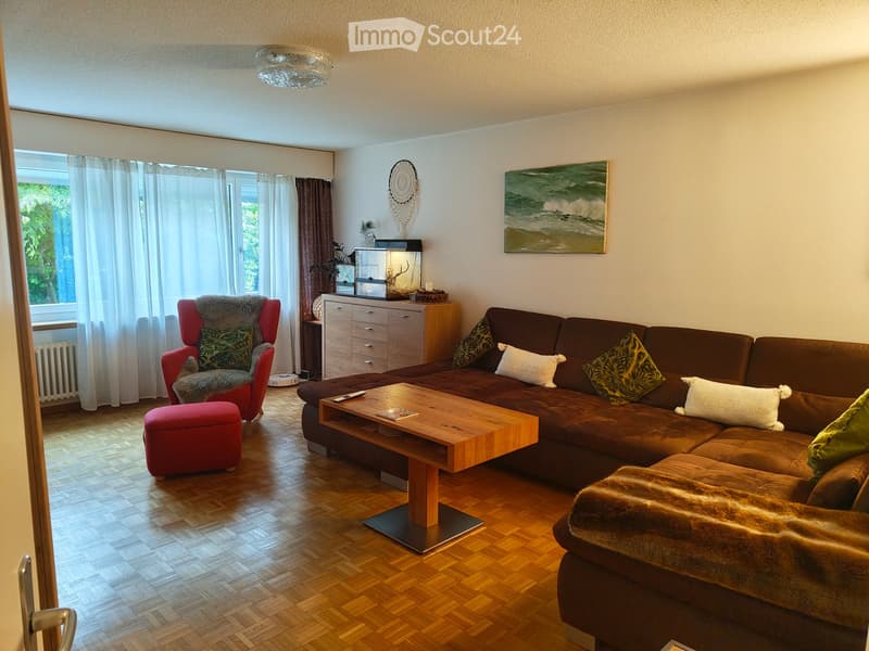 Bright living room with wooden flooring, large window with curtains, brown sectional sofa, wooden coffee table, red armchair with fur throw, ottoman, sideboard, and decorative items.