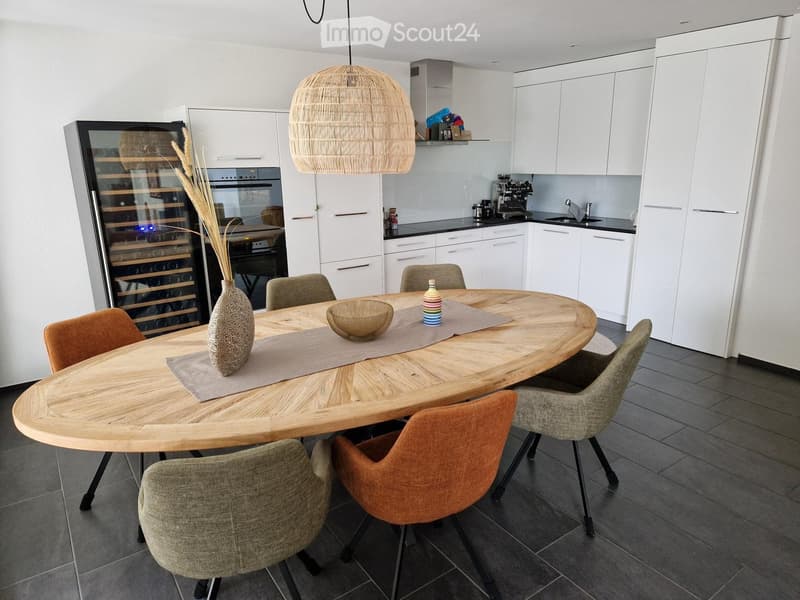 Modern kitchen with a round wooden dining table, six chairs, a wine cooler, and a coffee machine. The space features white cabinets, a hanging woven pendant light, and a sleek countertop.