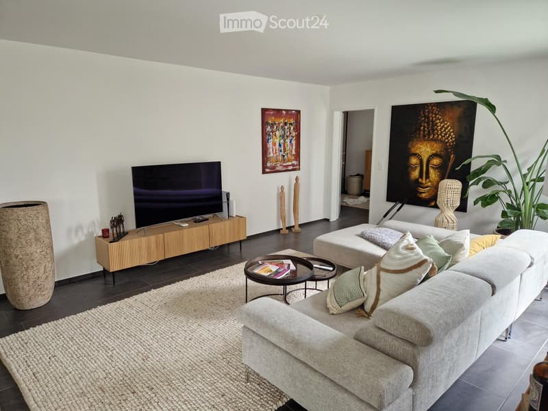 Modern living room with a grey sectional sofa, coffee table, TV, sideboard, and decorative plant. The space features a large, open layout with white walls and dark wooden flooring.