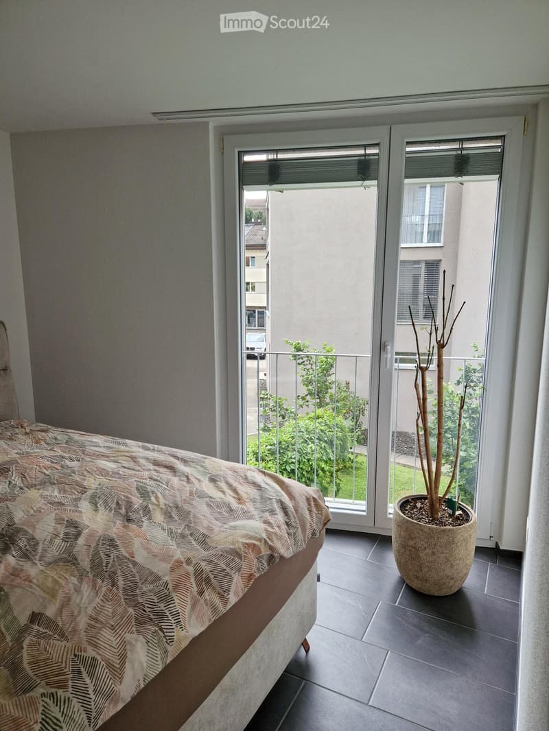 Bedroom with a bed covered in a patterned duvet, a large potted plant near the balcony entrance, and a view of a green outdoor area through large windows and a sliding door.