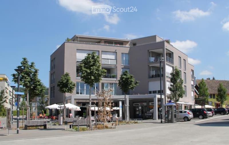 Modern multi-story building with balconies, large windows, and a ground-floor commercial area with outdoor seating.