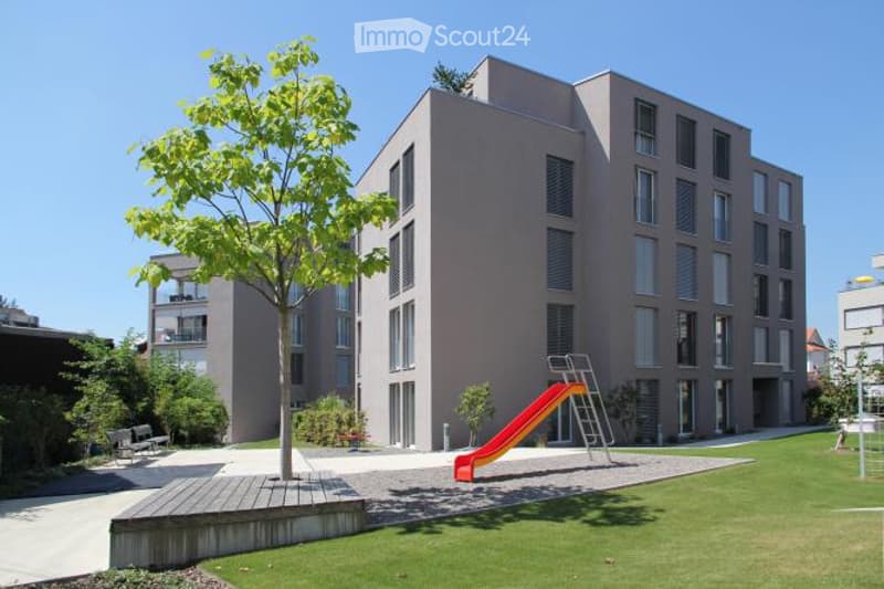 Modern multi-story residential building with large windows and balconies, surrounded by a well-maintained lawn, a small playground with a slide, and a few trees.