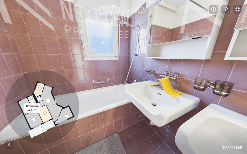 Bathroom with a bathtub, sink, mirror, and a yellow towel on the sink. The walls are tiled in a warm, reddish-brown color, and there is a window providing natural light.