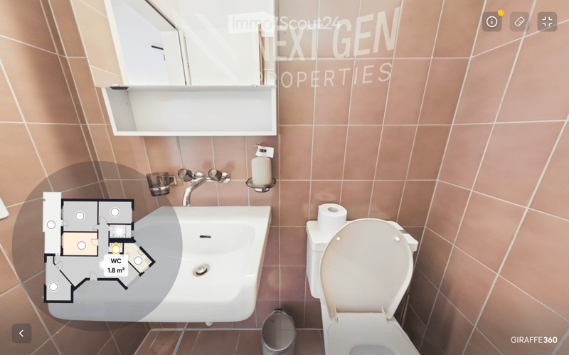 Modern bathroom with a white sink, chrome faucet, toilet, and a soap dispenser. The walls are tiled in a warm, peach-colored pattern, and a mirror is mounted above the sink. A small shelf is also visible.