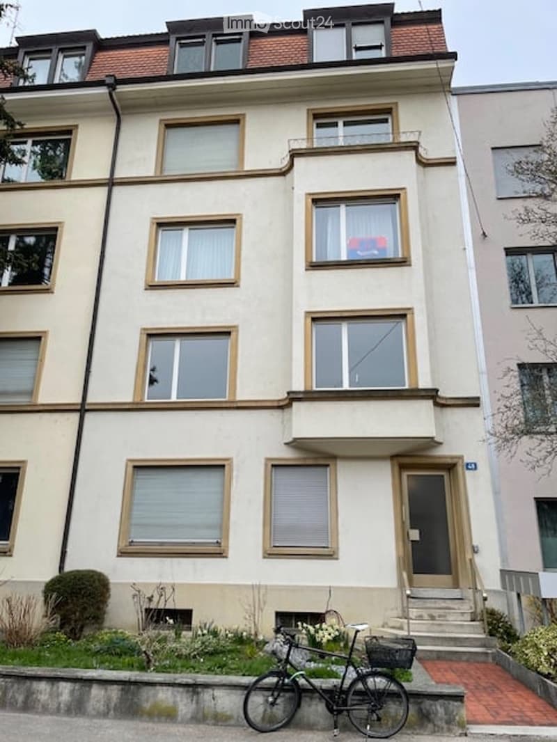 Three-story apartment building with white exterior, multiple windows, two bicycles parked in front, a small garden area with plants in front of the building.