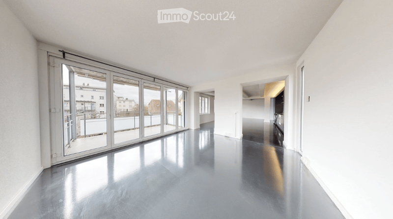 Empty room with large sliding glass doors leading to a balcony, polished concrete floor, white walls, and visible kitchen area in the background.