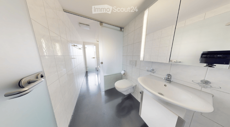 Modern bathroom with white tiled walls, a toilet, a sink with a mirror, and a cabinet. The floor is dark grey, and the space is well-lit with natural light coming from the left side.