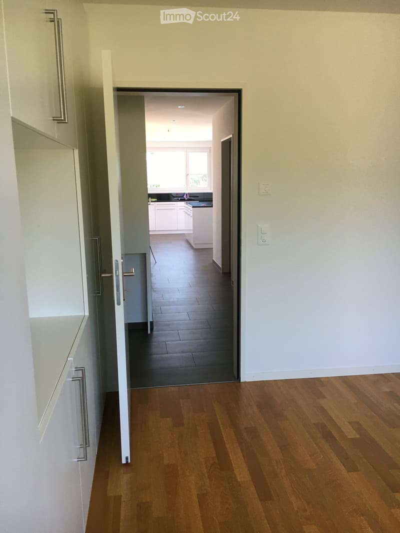 A clean, modern hallway with wooden flooring, white walls, and a door leading to a kitchen area with white cabinets and a dark countertop.