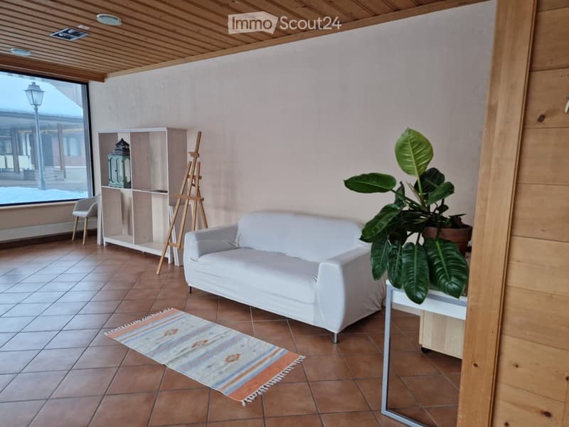 A modern living room with a white sofa, a small table with a large leafy plant, a shelf with decorative items, a small chair, and an easel near the window.