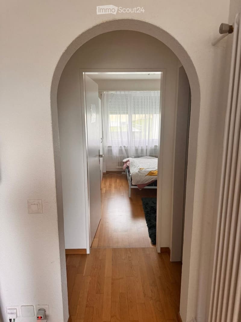 A narrow hallway with wooden flooring and light-colored walls, leading to a bedroom with a bed, rug, and window with curtains.