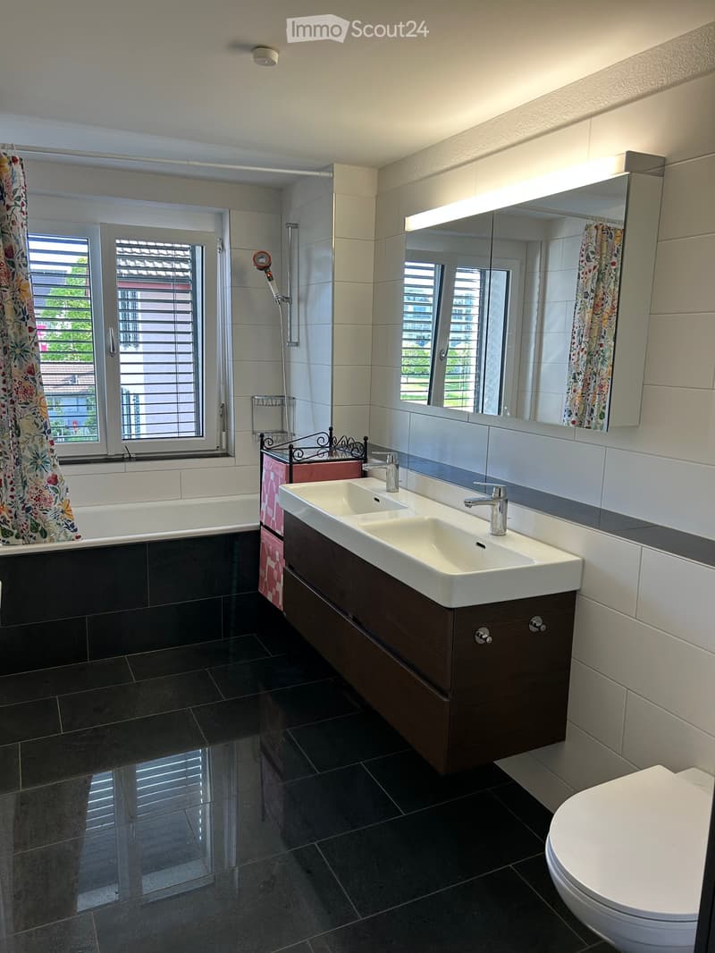 Modern bathroom with dark tiled floor, white sink with dual faucets, large mirror, bathtub with floral shower curtain, and toilet.