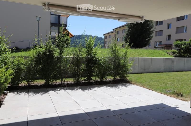 A well-maintained garden area with a tiled patio, neatly trimmed bushes, and a low concrete wall separating the space from a grassy lawn. The background shows a residential building and a clear blue sky.