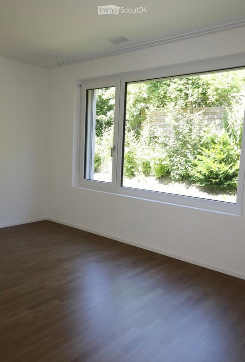 Empty room with wooden flooring and white walls, featuring large windows with white frames that open to a garden view.