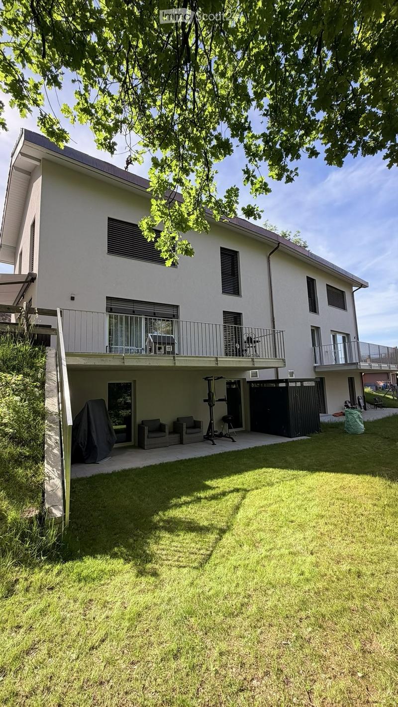 A modern two-story house with white exterior, multiple windows, balconies, and a well-maintained lawn. Outdoor furniture and exercise equipment are visible on the patio.