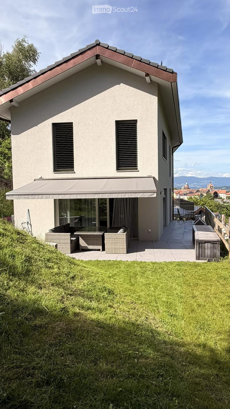 Two-story house with a modern design, featuring a covered patio area with outdoor furniture, a grassy lawn, and a scenic view of a town and mountains in the background.