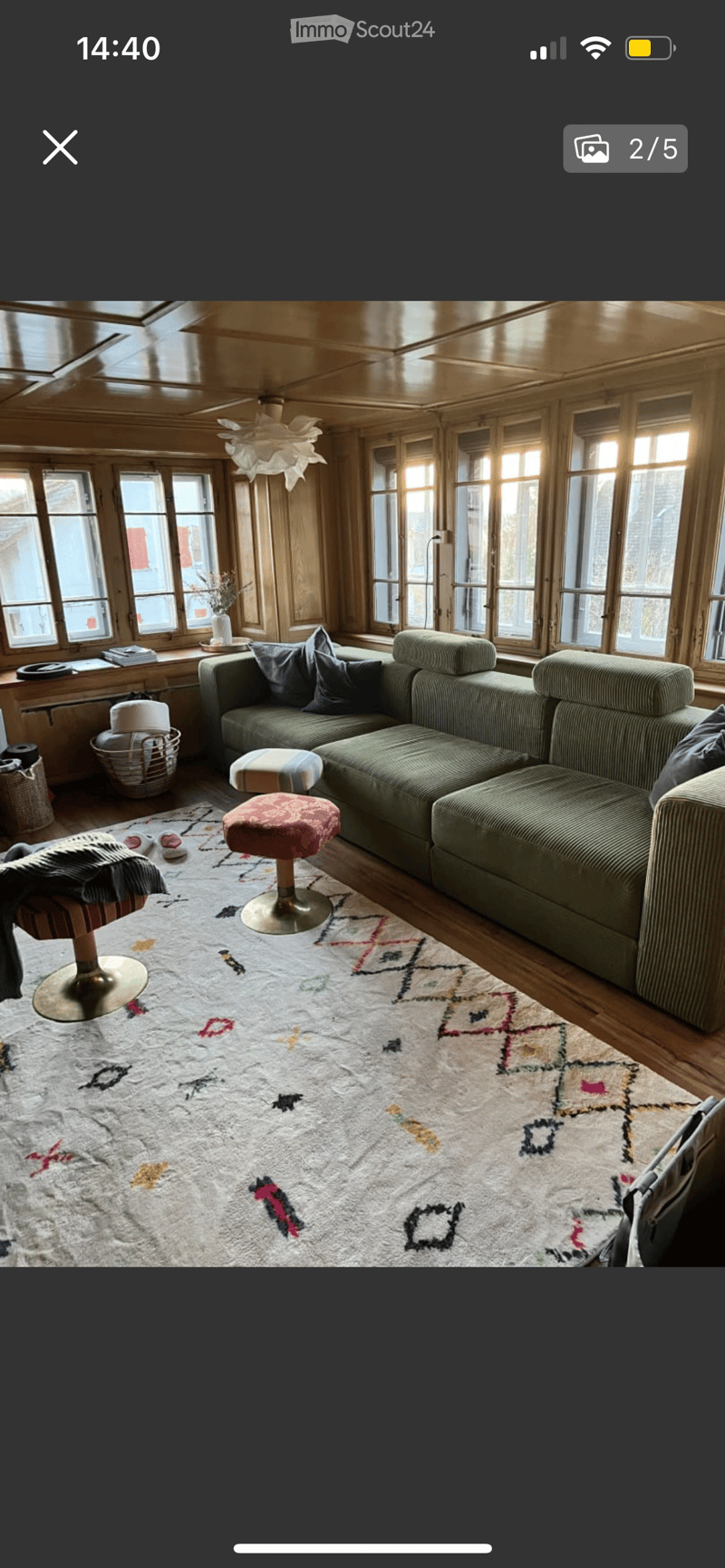 A cozy living room with wooden walls and large windows, featuring a green sectional sofa, colorful patterned rug, and various seating options including a stool and chair.