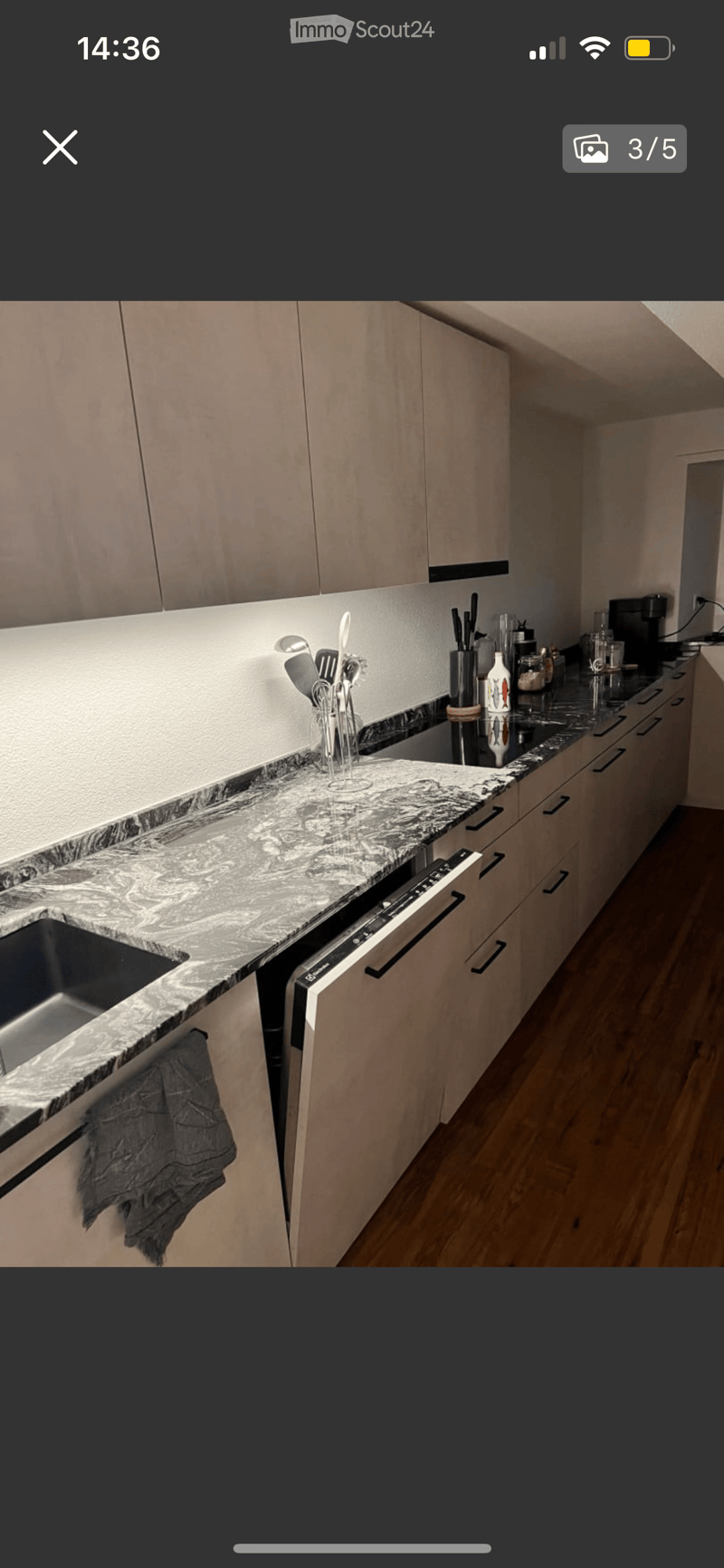 Modern kitchen with light wood cabinets, marble countertop, stainless steel sink, dishwasher, and various utensils and appliances on the counter.