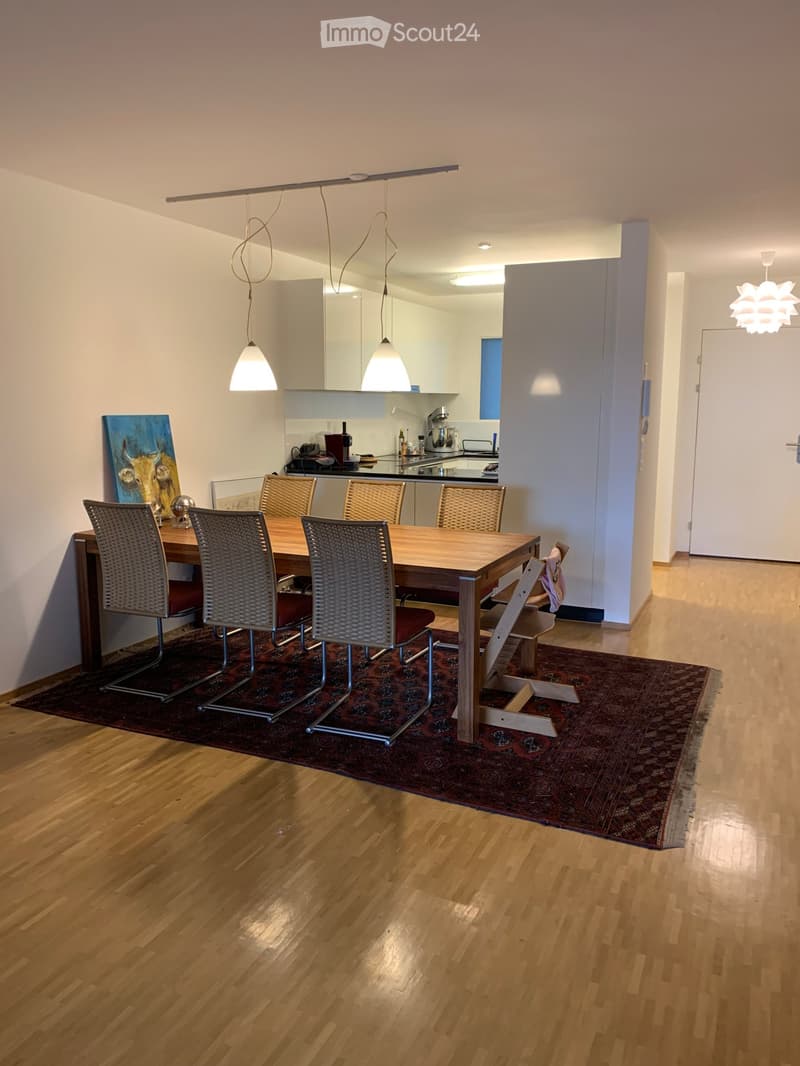 Dining area with a wooden dining table and six chairs, pendant lights, a kitchen area with appliances, and a painting on the wall.