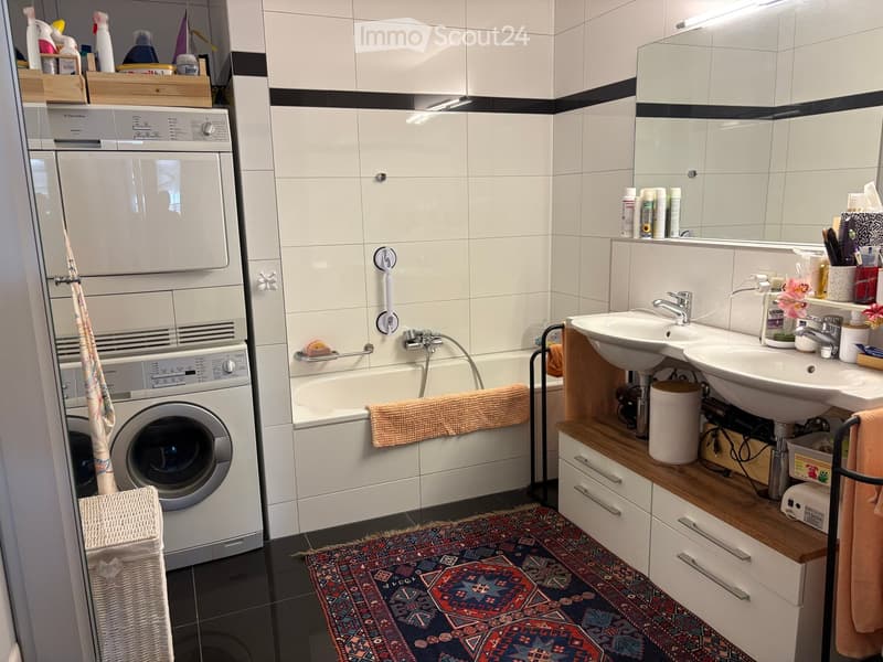 Modern bathroom with white tiles, a bathtub, two sinks, a washing machine, and a wicker basket. Features include a shower area with a curtain, a towel on the edge of the tub, and a colorful rug on the floor.
