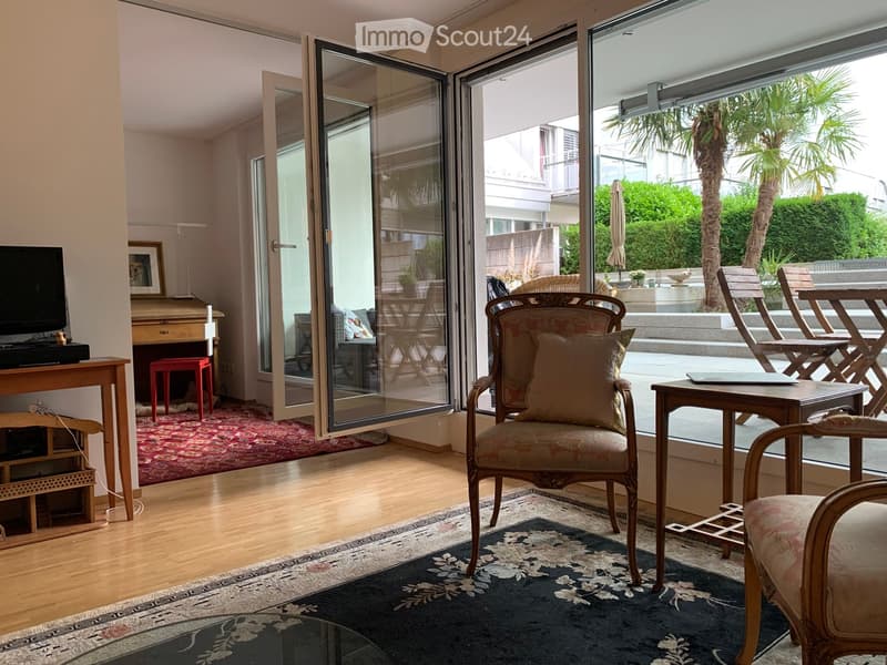 Bright living room with large glass doors opening to a patio, featuring a patterned rug, wooden furniture, a piano, and a television on a wooden stand.