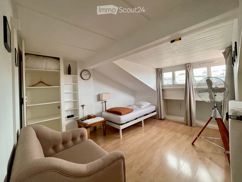 Cozy attic bedroom with wooden floors, a single bed, a sofa, a small table, a fan, shelves, and a clock on the wall.