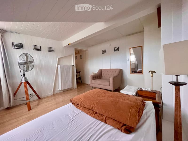 Cozy bedroom with wooden floors, white paneled walls, a bed with orange bedding, a pink armchair, a standing fan, a mirror, and a small table with a lamp.