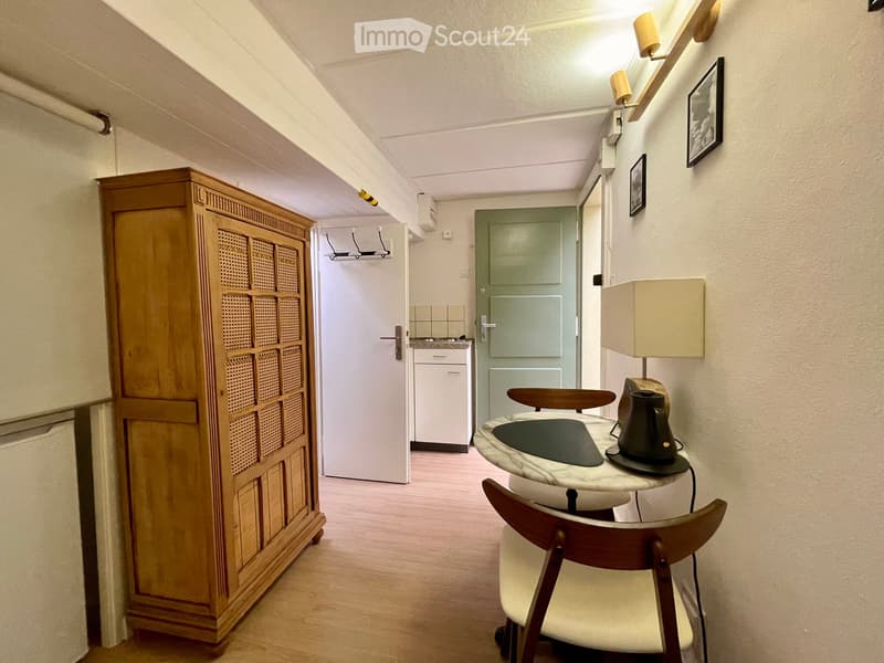 A small, furnished room with a wooden floor, a round table with a marble top, a chair, a lamp, a wooden cabinet, and a small kitchen area with a refrigerator and stove visible through an open door.