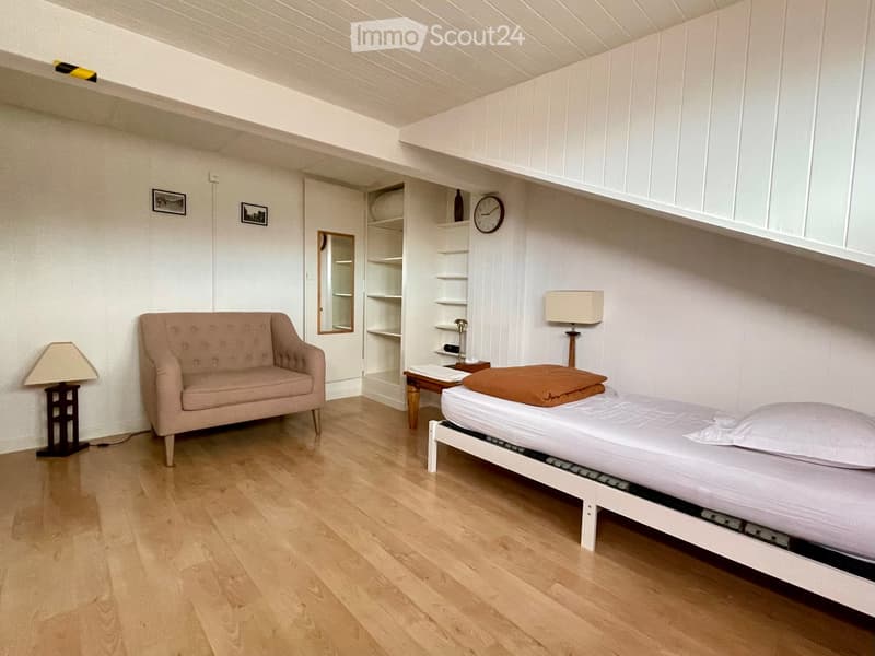 Cozy, well-lit room with a single bed, sofa, small table, and shelves. The room features wooden flooring and white walls.